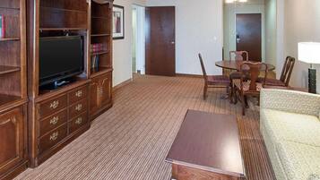 Habitación de hotel con mueble de madera para entretenimiento, televisión de pantalla plana, mesa de comedor con sillas, sofá y ventilador de techo.