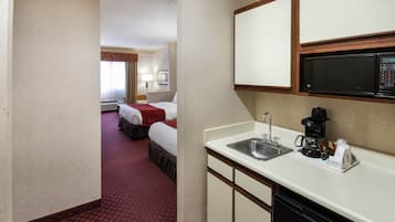 Habitación de hotel con kitchenette, que incluye microondas, pileta y cafetera. Tiene una cama con una manta roja y una ventana con cortinas.