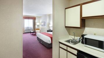 A hotel room with a bed, a desk, a chair, a window with curtains, a wall-mounted TV, and a kitchenette with a microwave and sink.