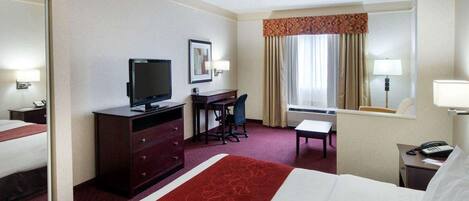 A hotel room with a large bed, a desk with a chair, a television, a small table, and a window with curtains.