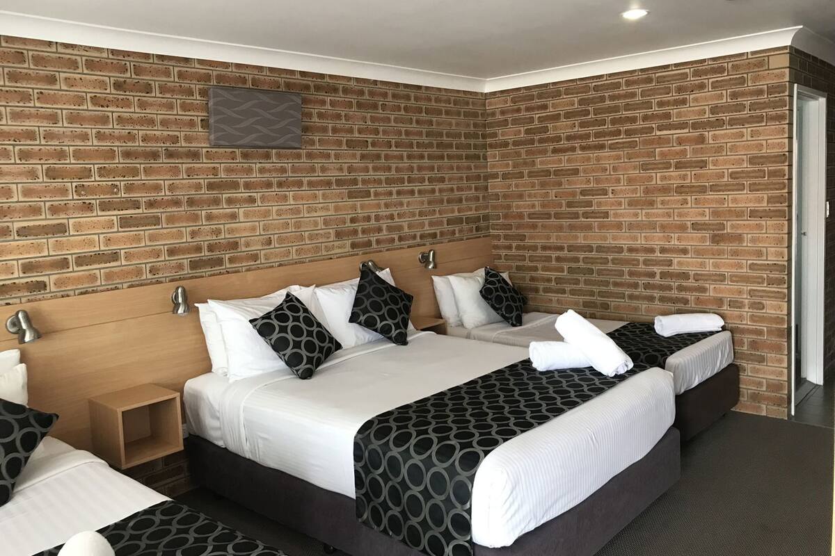 A hotel room with a brick wall, two beds with patterned bedding, a wooden headboard, and a small bedside table.