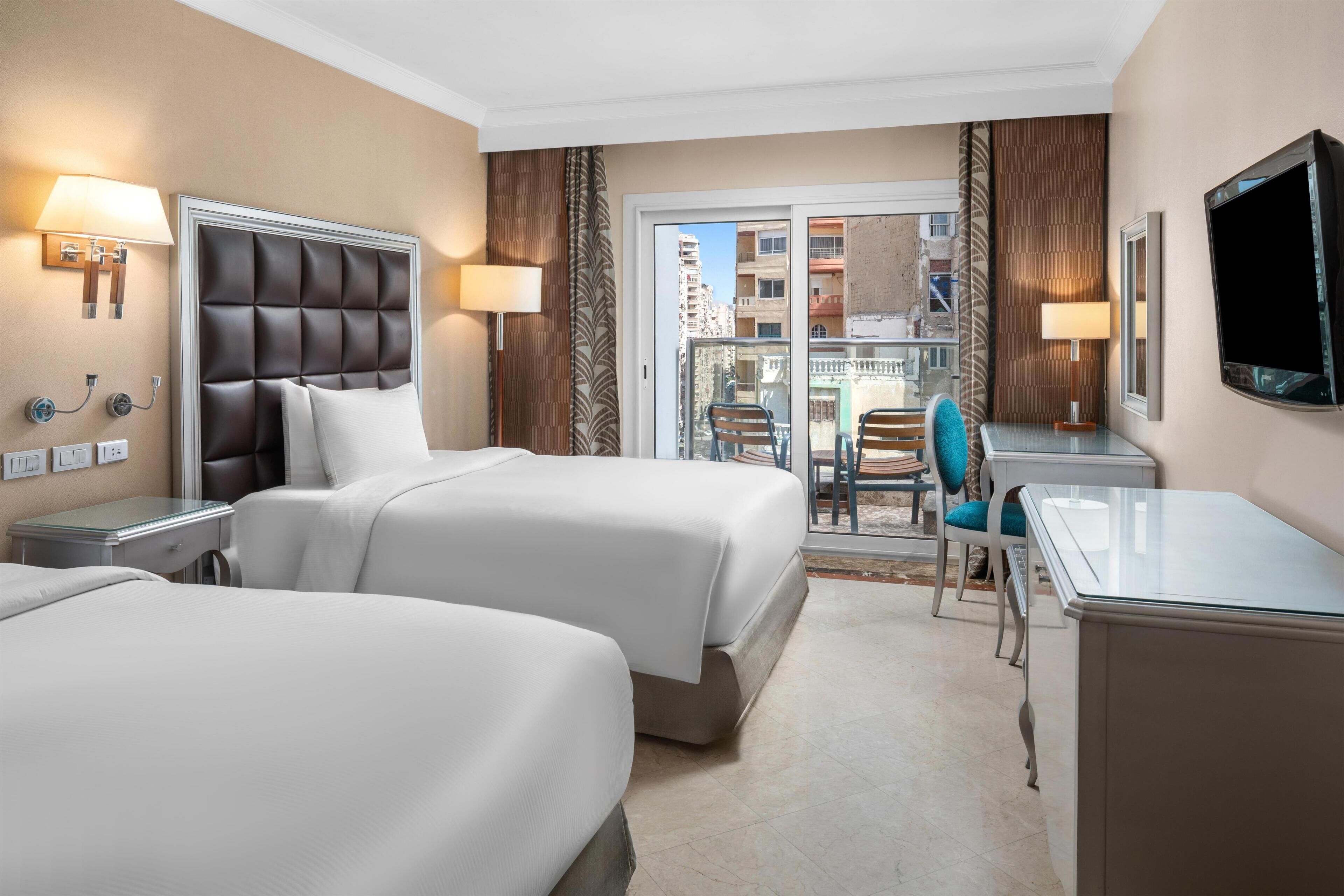 A hotel room with two beds, a desk, a chair, and a TV.