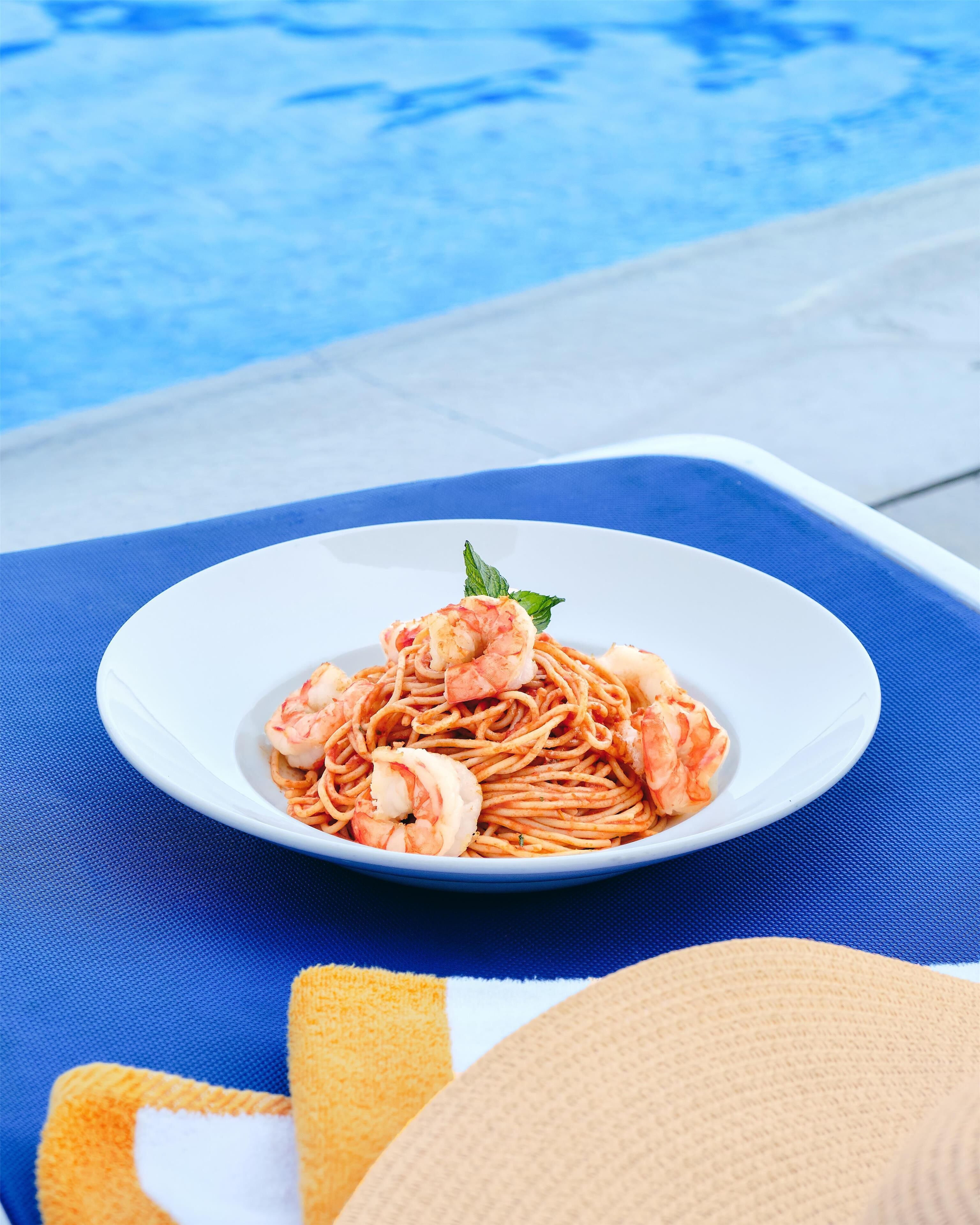 Une assiette de pâtes aux crevettes, servie dans une assiette blanche, posée sur une chaise bleue près d’une piscine.