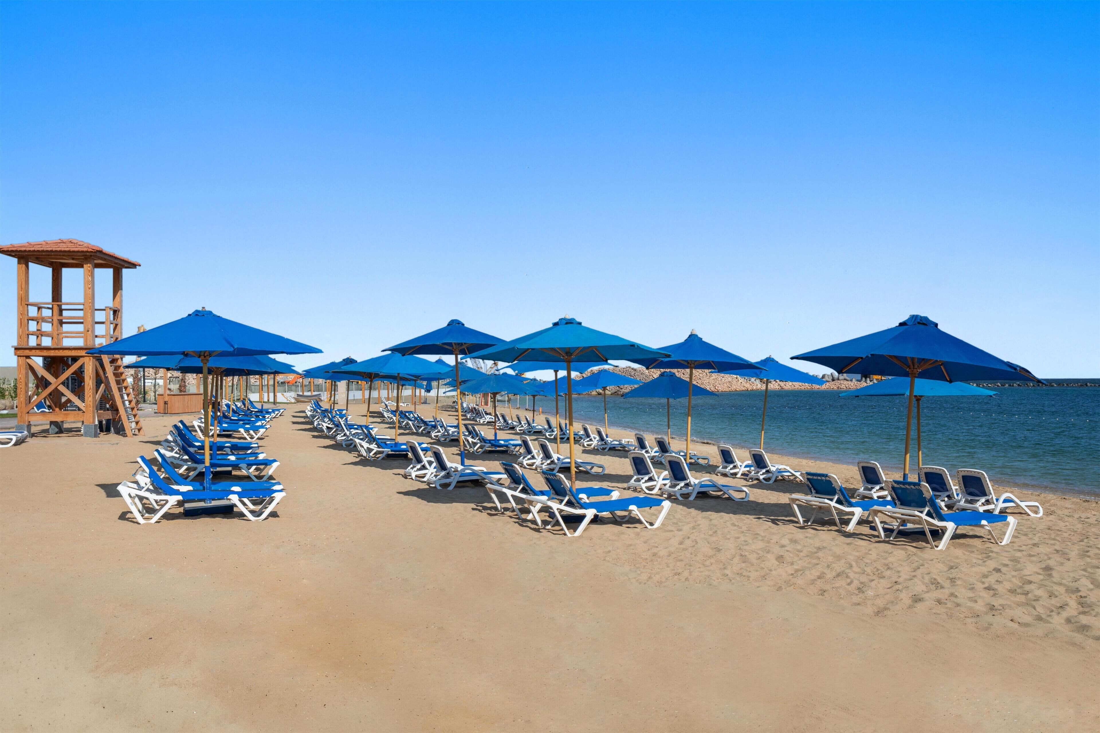 Ein Strand mit Reihen von Liegestühlen und blauen Sonnenschirmen, einer Holzkonstruktion und einem klaren Himmel.