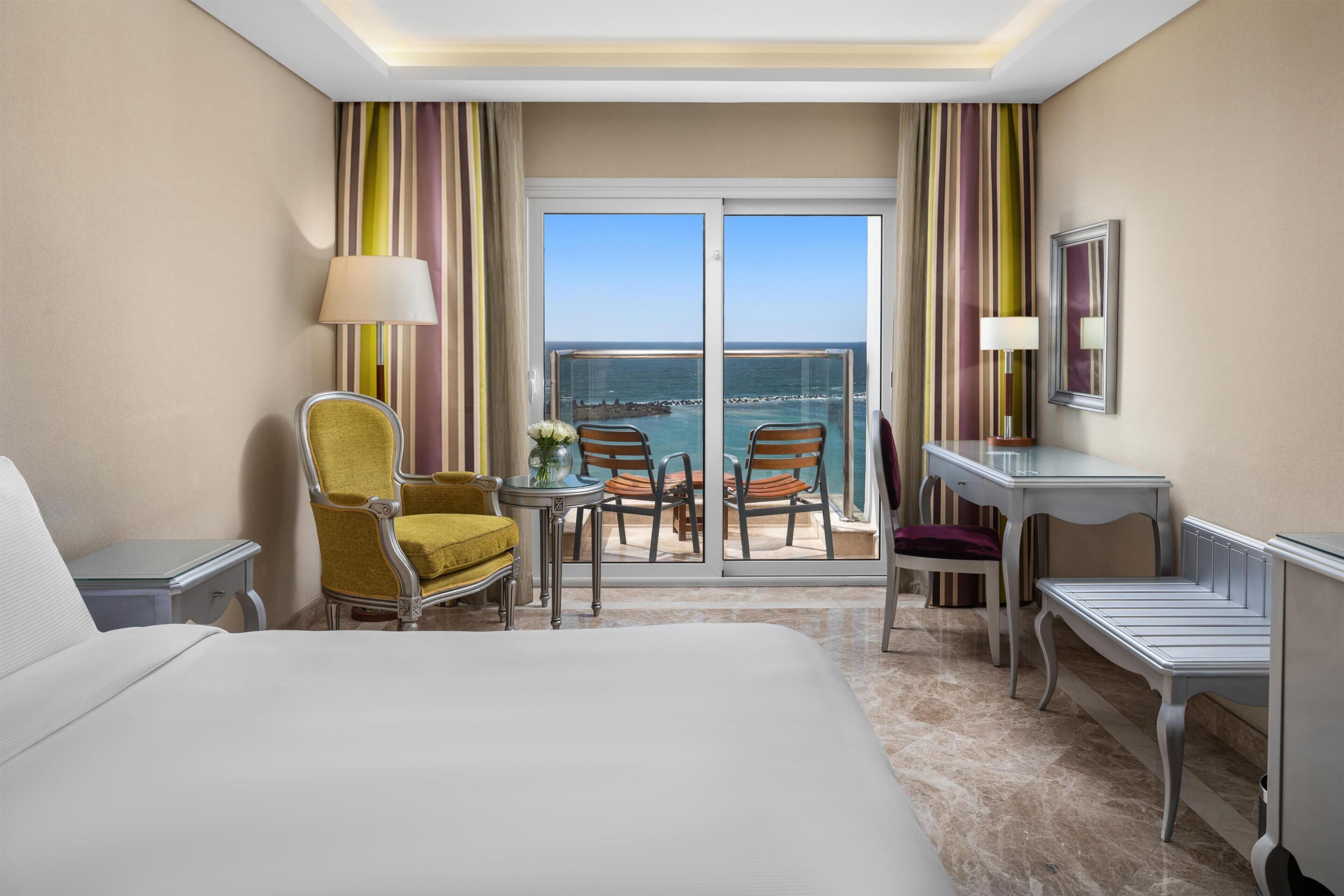 Een hotelkamer met een bed, twee stoelen, een bureau, een lamp en uitzicht op zee vanaf een balkon.