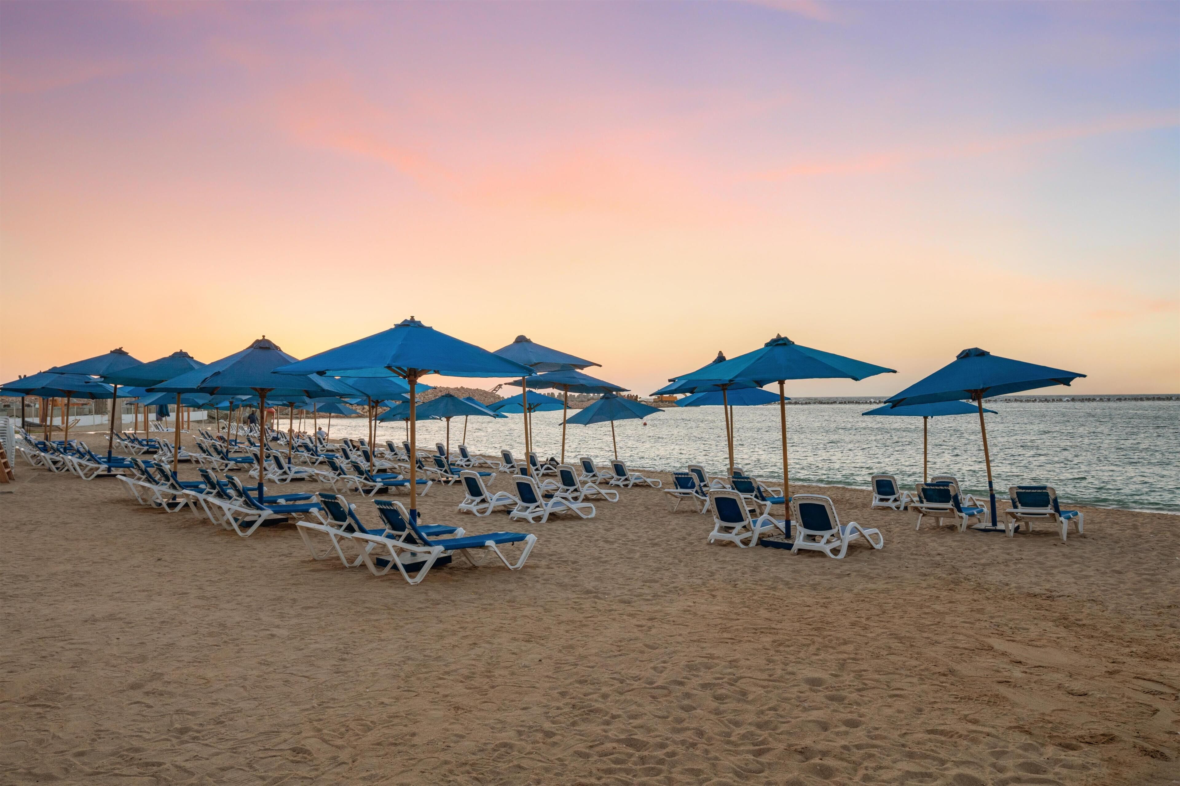 A beach with rows of sun loungers and blue umbrellas.