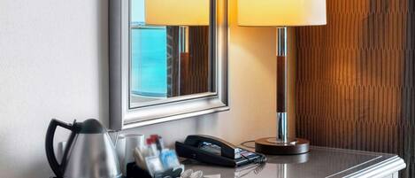 Habitación de hotel con un escritorio de madera pulida, un espejo que refleja dos lámparas, una tetera, un teléfono y una bandeja con artículos de aseo.