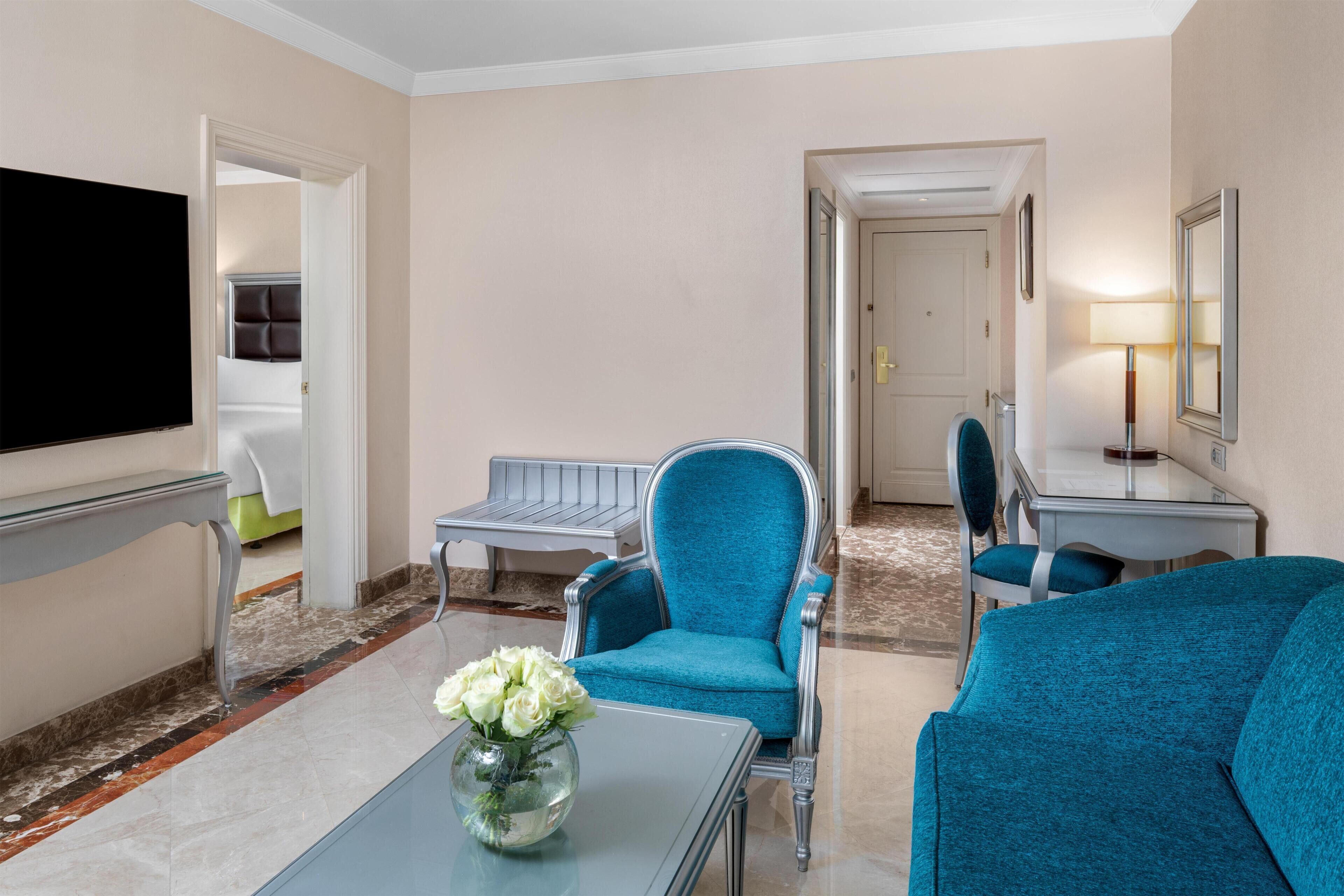 Quarto de hotel com televisão de tela plana, sofá, poltrona, mesa pequena com um vaso de flores e uma cama com cabeceira.