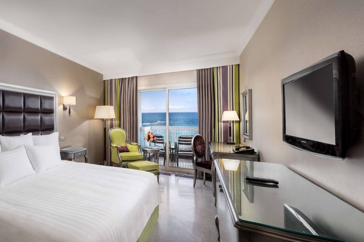 A hotel room with a large bed, a TV, a desk with a chair, a phone, and a view of the sea through a balcony.