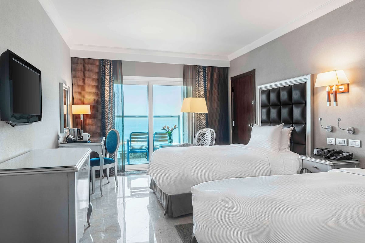 A hotel room with two beds, a desk, a chair, a TV, and a balcony with a view.