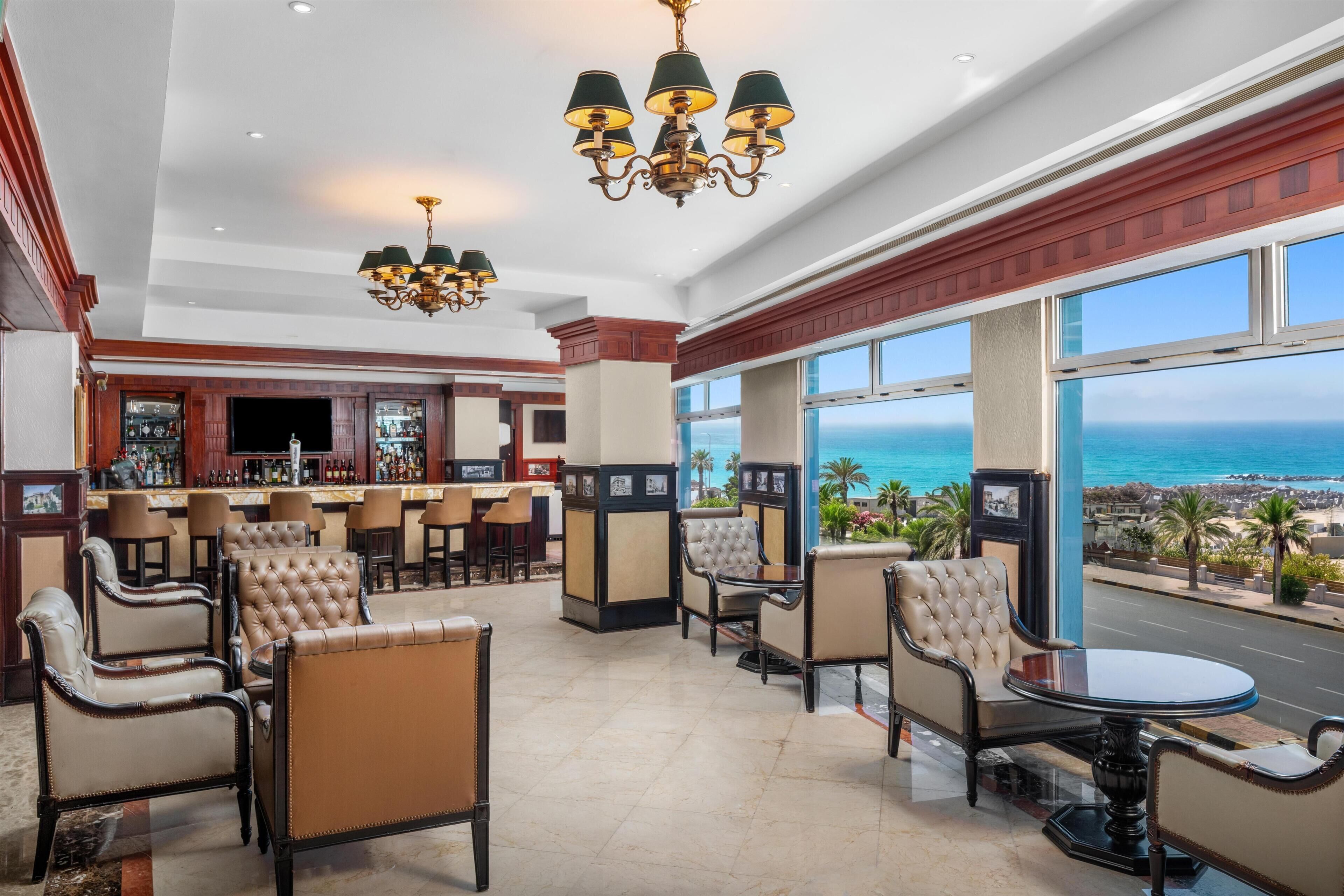 Un vestíbulo de hotel con zona de bar, asientos con sillones cómodos y grandes ventanales con vistas al océano.