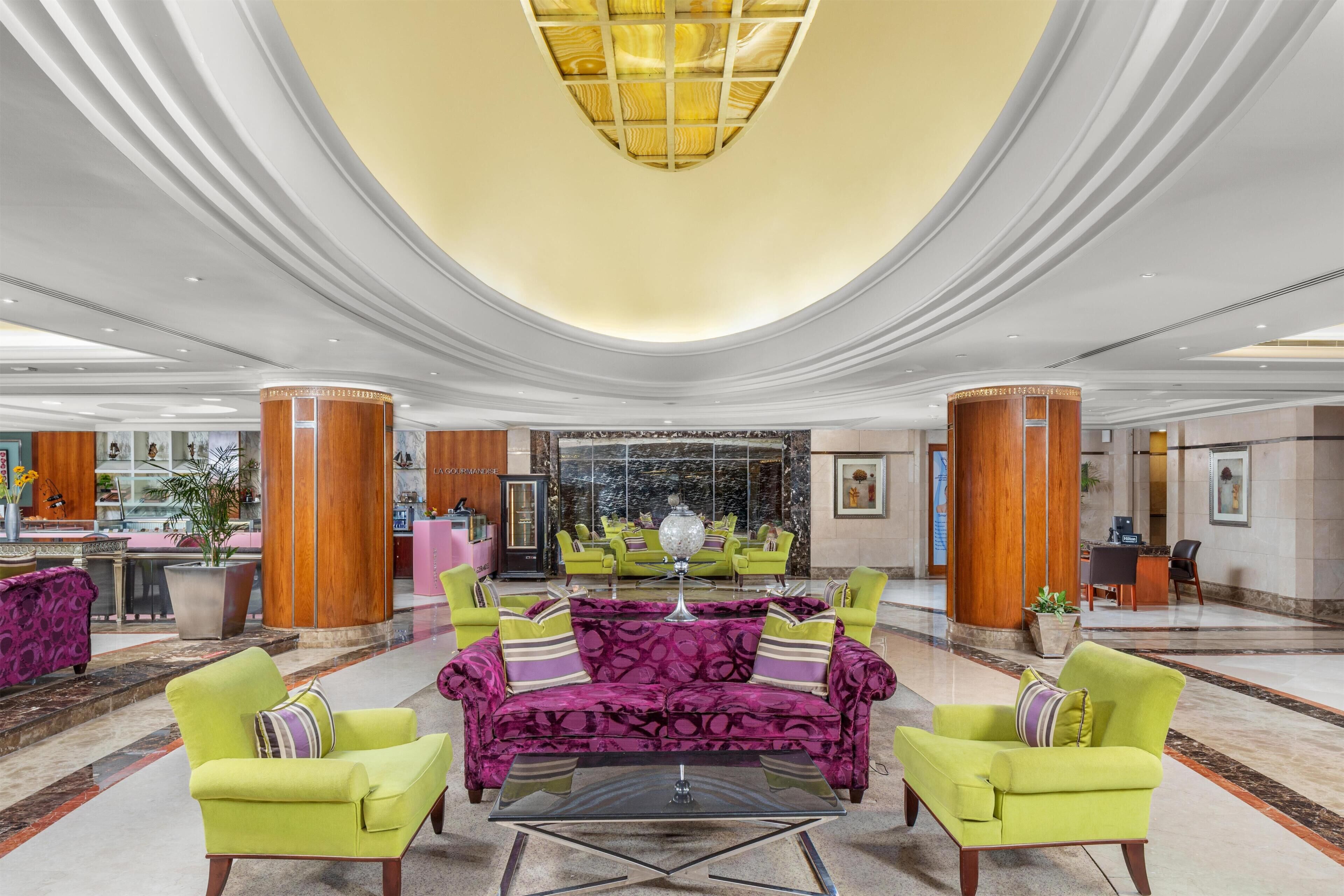 A spacious lobby with a central seating area featuring a purple sofa and green armchairs, a reception desk, and a large glass wall.
