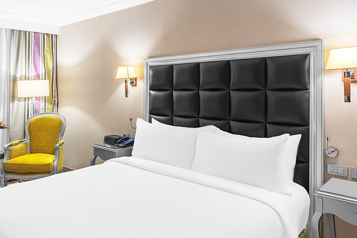 A hotel room with a large bed, a yellow armchair, a bedside table with a phone, and two wall-mounted lamps.
