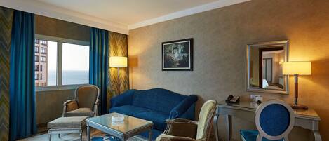 A hotel room with a blue sofa, two armchairs, a small table, a desk with a lamp, a telephone, and a painting on the wall.