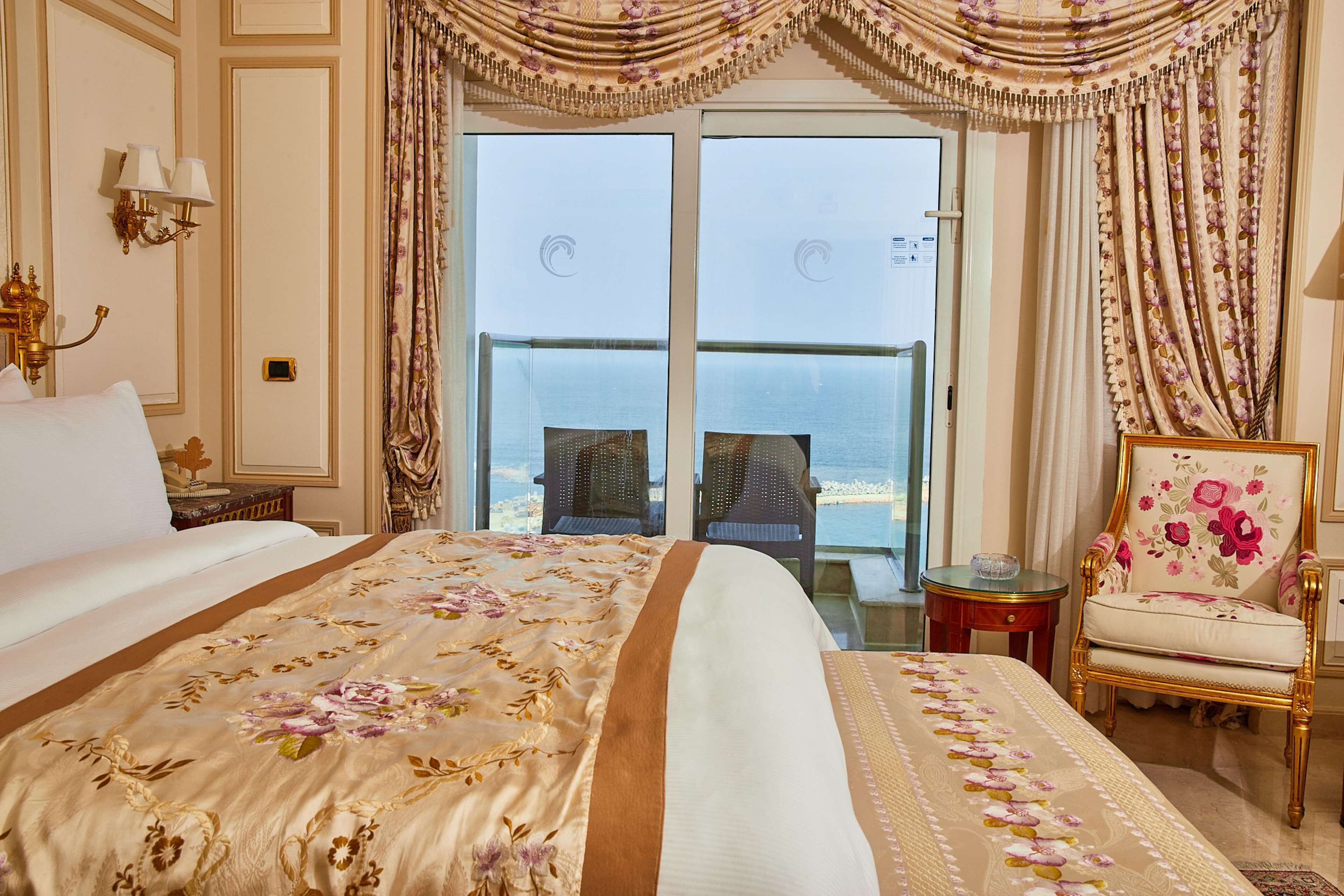 A hotel room with a large bed, a balcony with two chairs, and a floral chair.