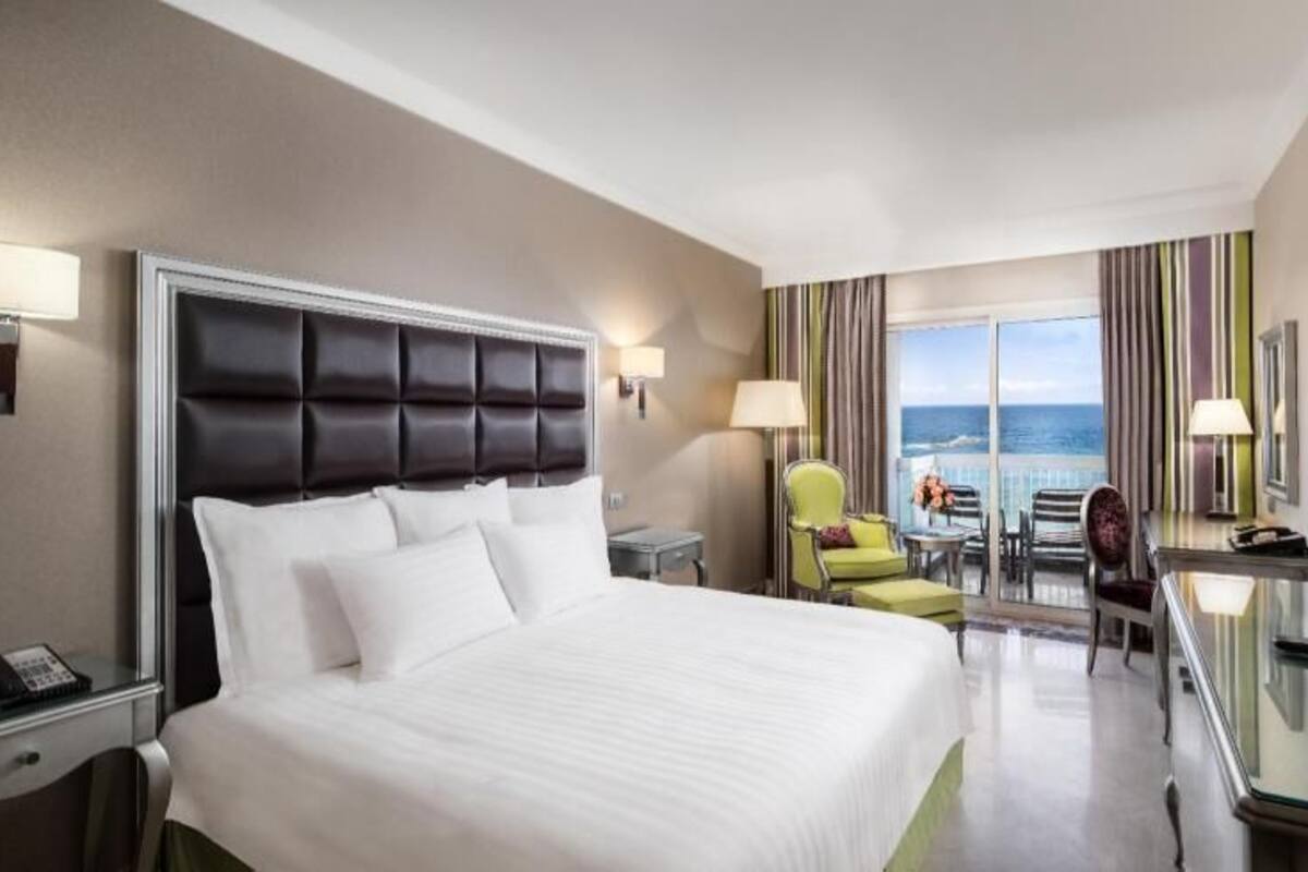 A hotel room with a large bed, a desk with a flat-screen TV, a chair, a small table, and a view of the ocean.