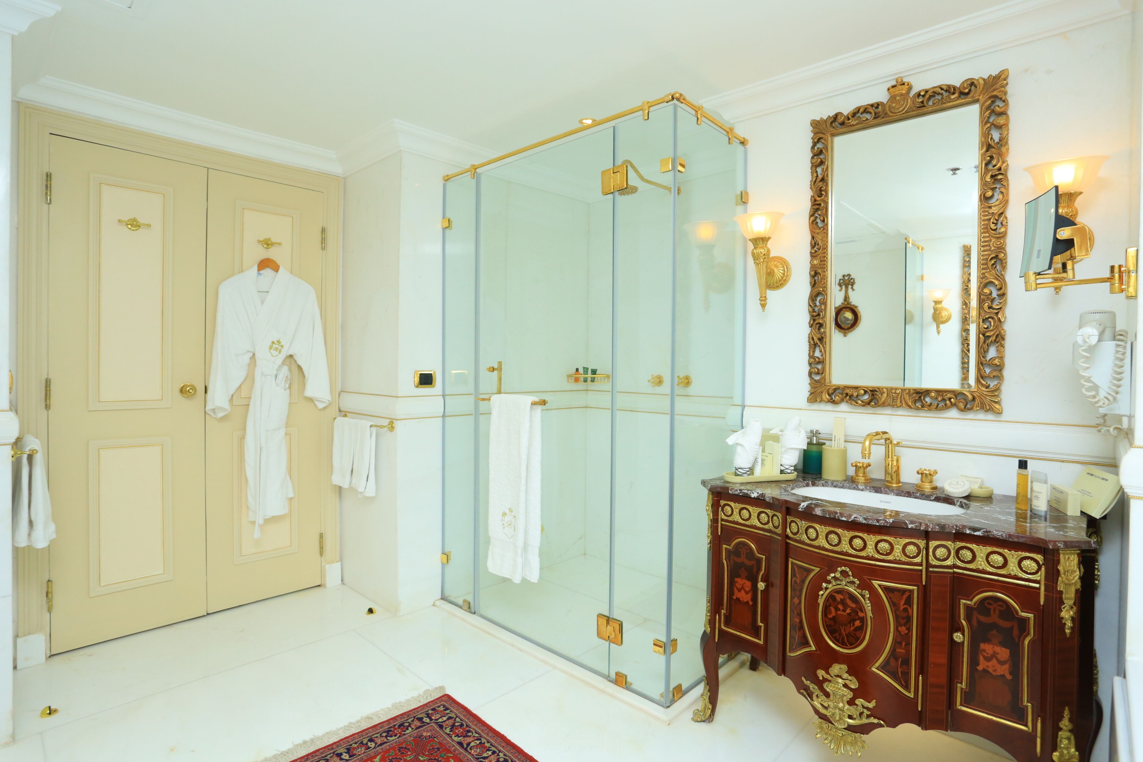 Een ruime badkamer met een douchecabine van glas, een wastafel met gedetailleerd houtwerk en een grote spiegel met een sierlijk omlijsting.