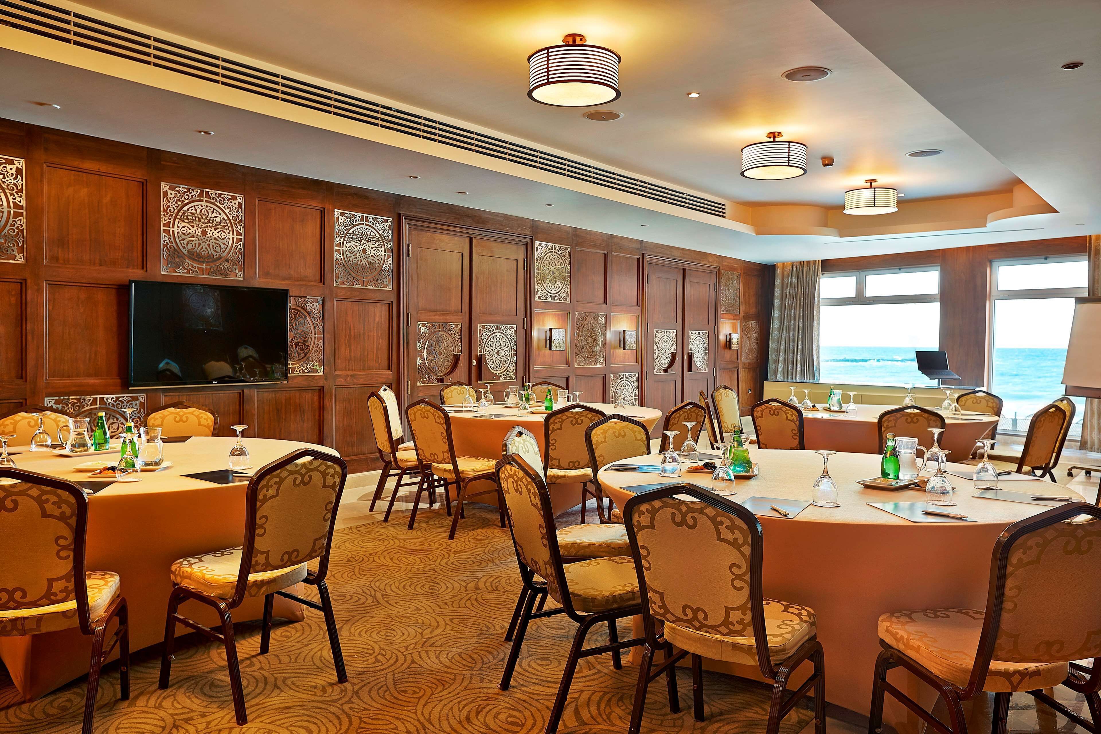 Una espaciosa sala de conferencias con varias mesas y sillas, amplias ventanas con vista al océano y paneles de madera decorativos.
