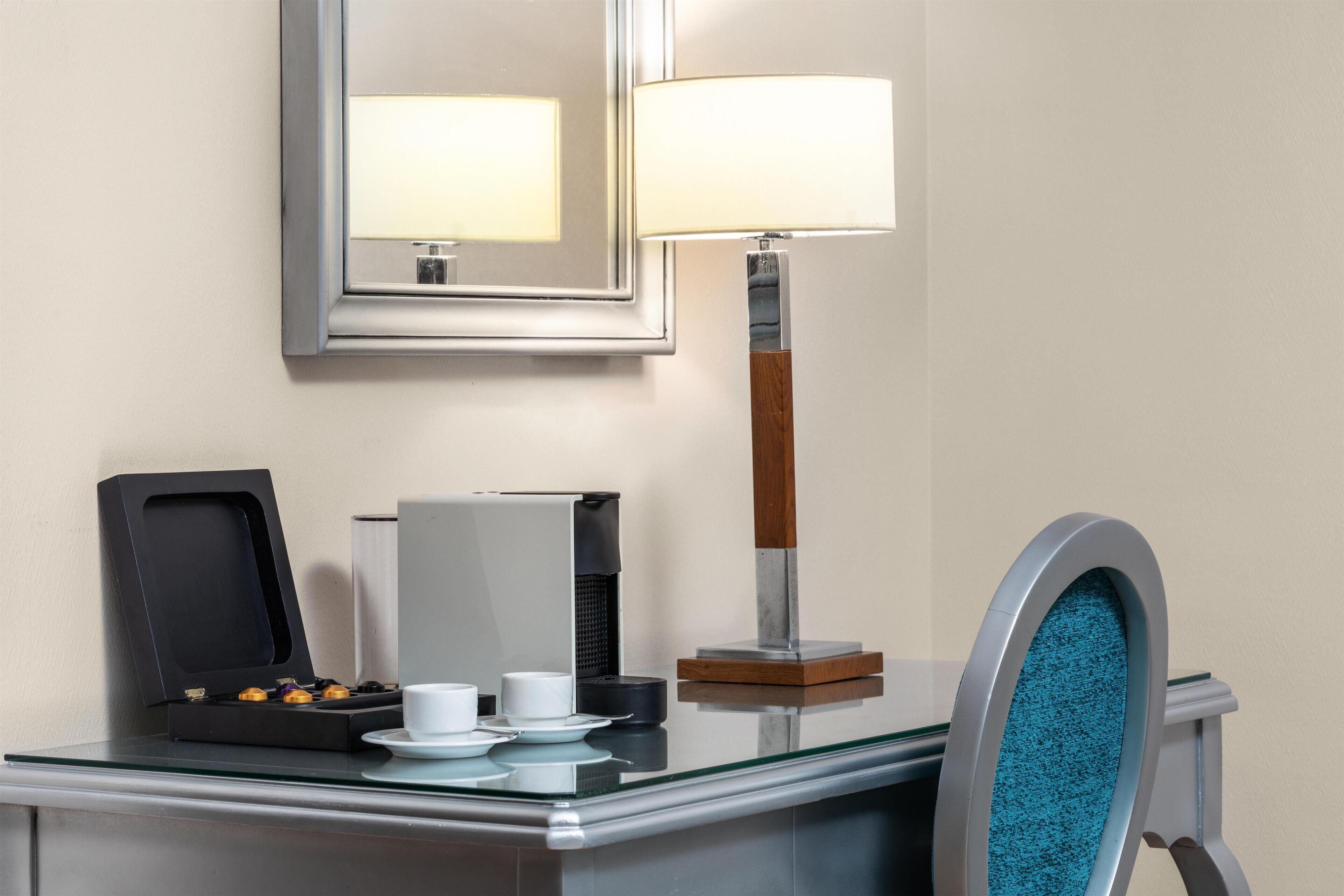 A hotel room with a glass table, a coffee maker, a tea set, a mirror, and a lamp.