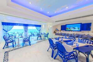 Un moderno comedor con sillas y mesas de cuadros azules, una pantalla de televisión grande y vistas al mar a través de amplios ventanales.