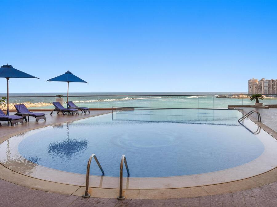 Un área de piscina con sillones de playa y sombrillas, con vista a una playa y al océano.