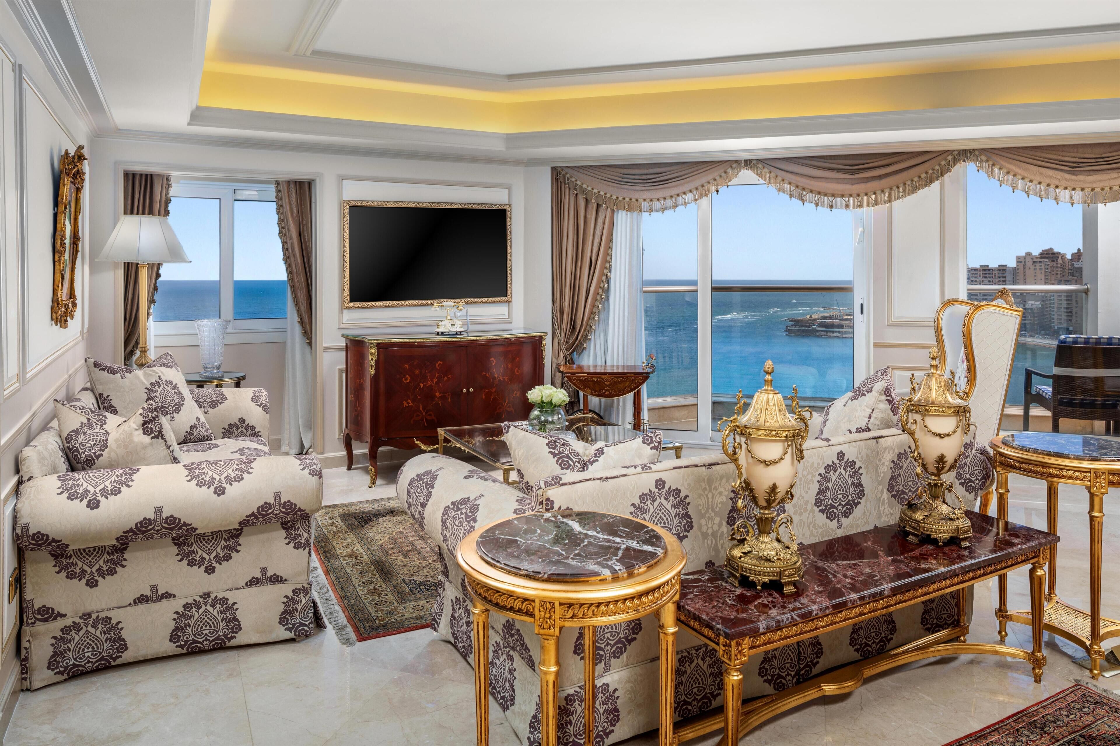Ein luxuriöses Wohnzimmer mit einer Couch, einem Couchtisch und einem Fernseher. Durch große Fenster ist der Meerblick zu sehen.