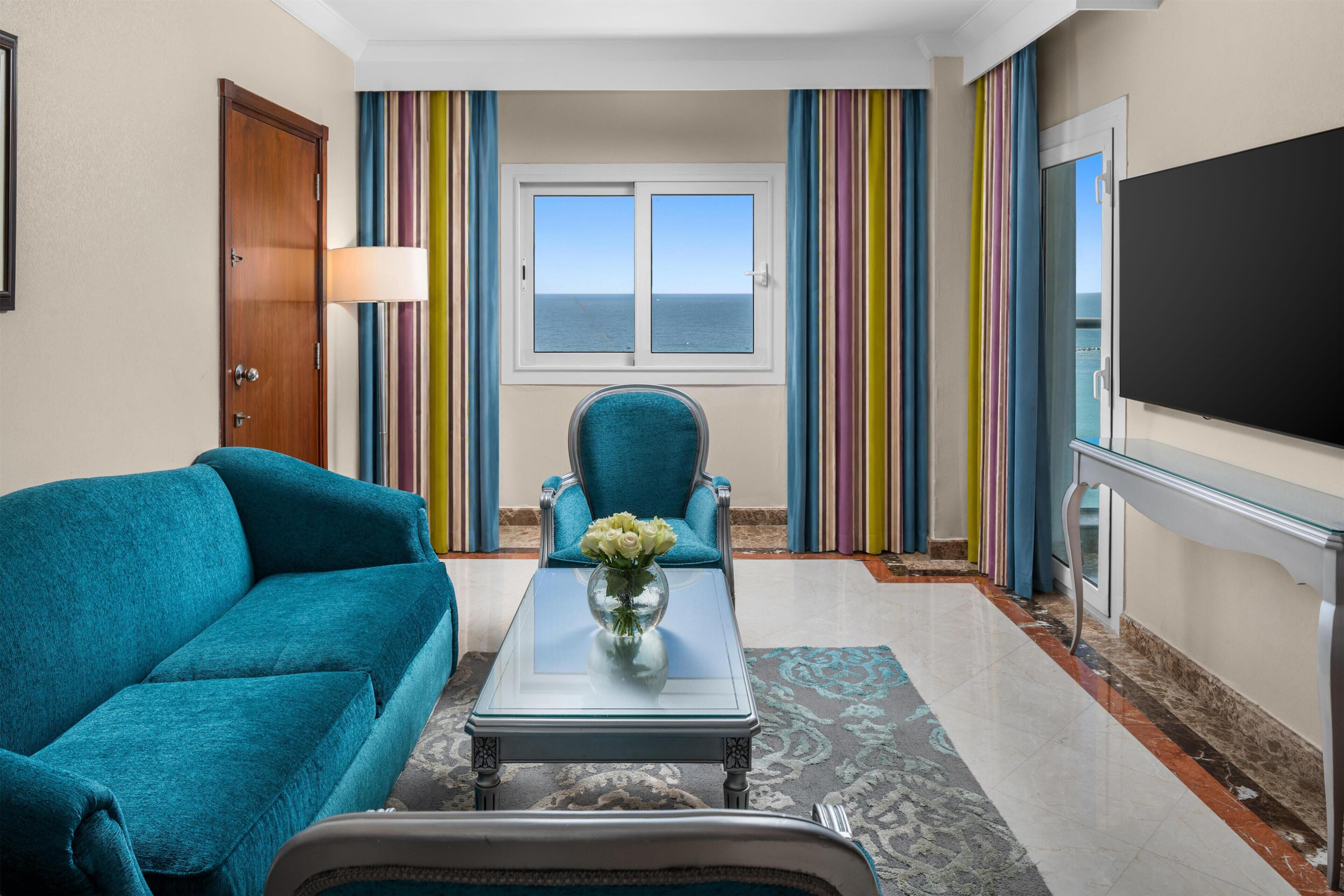 Ein Zimmer mit einem türkisfarbenen Sofa, einem Glastisch mit Blumenvase, einem blauen Sessel, einem großen Fernseher und Meerblick durch ein Fenster mit gestreiften Vorhängen.