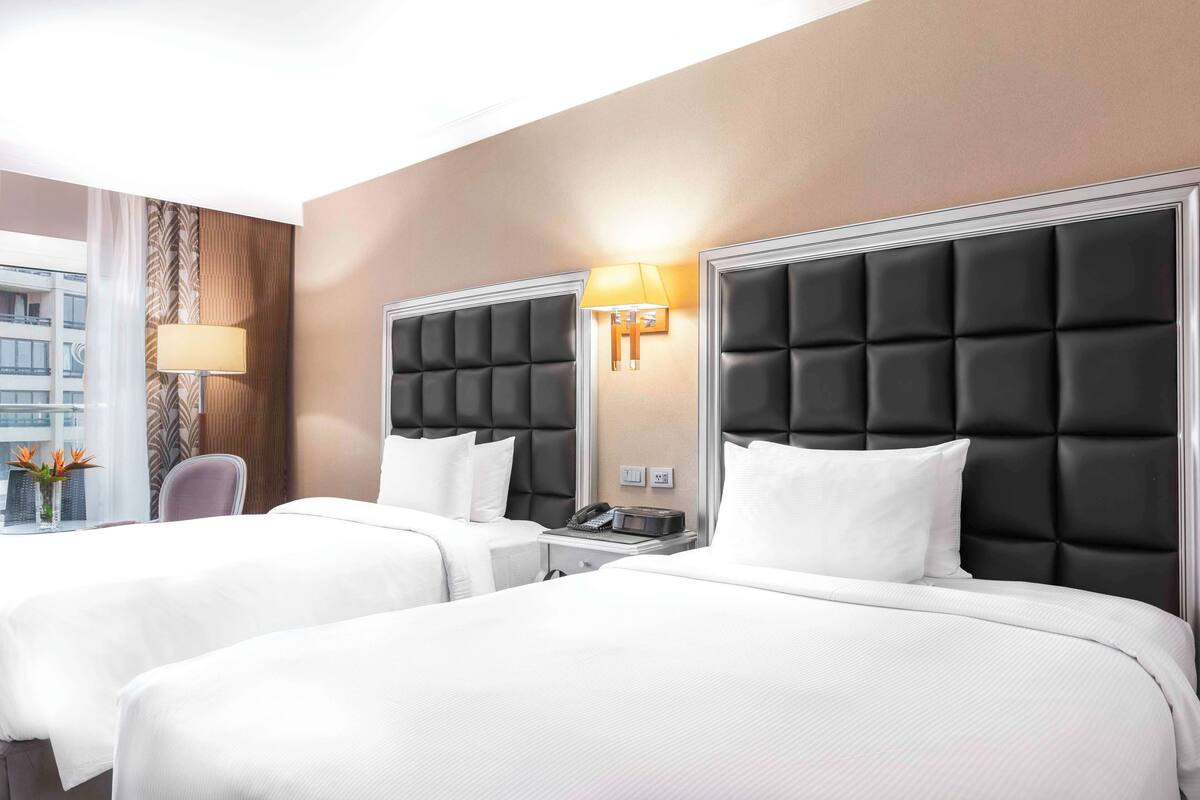 A hotel room with two beds, a bedside table with a lamp, a chair, and a window with a view of buildings.