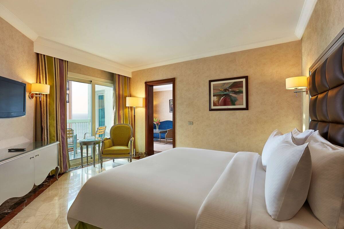 A hotel room with a large bed, a desk, a chair, a TV, and a balcony with a view.