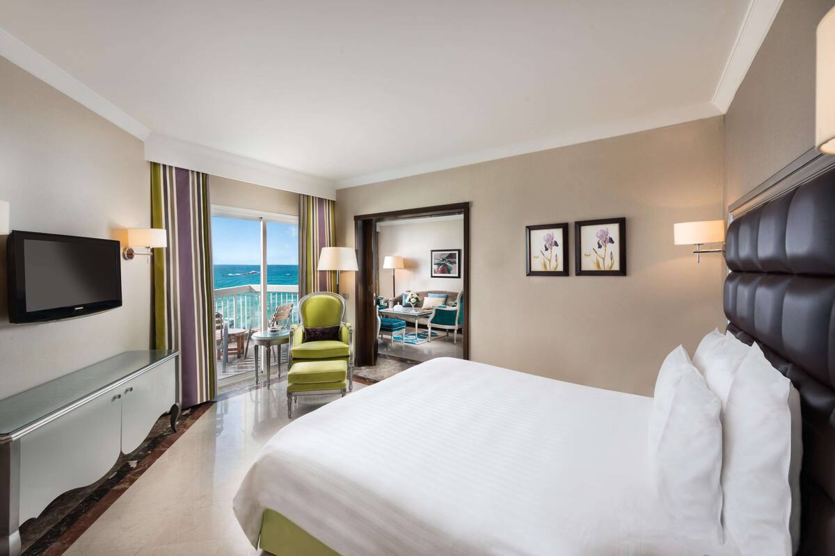 A hotel room with a large bed, a television, a desk, and a chair. There is a balcony with a view of the ocean.