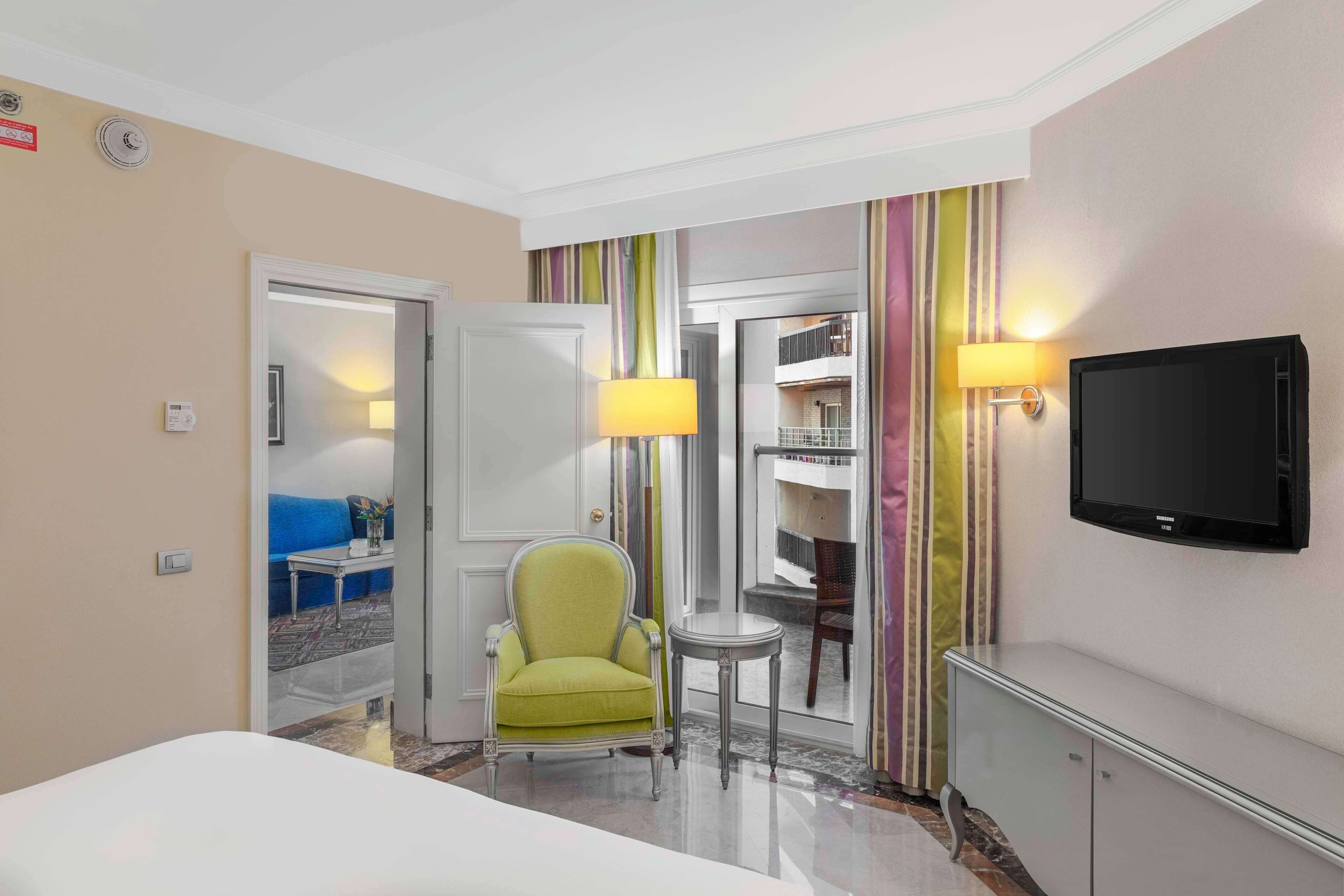 A hotel room with a bed, a green armchair, a small table, a TV, and a door leading to a balcony.