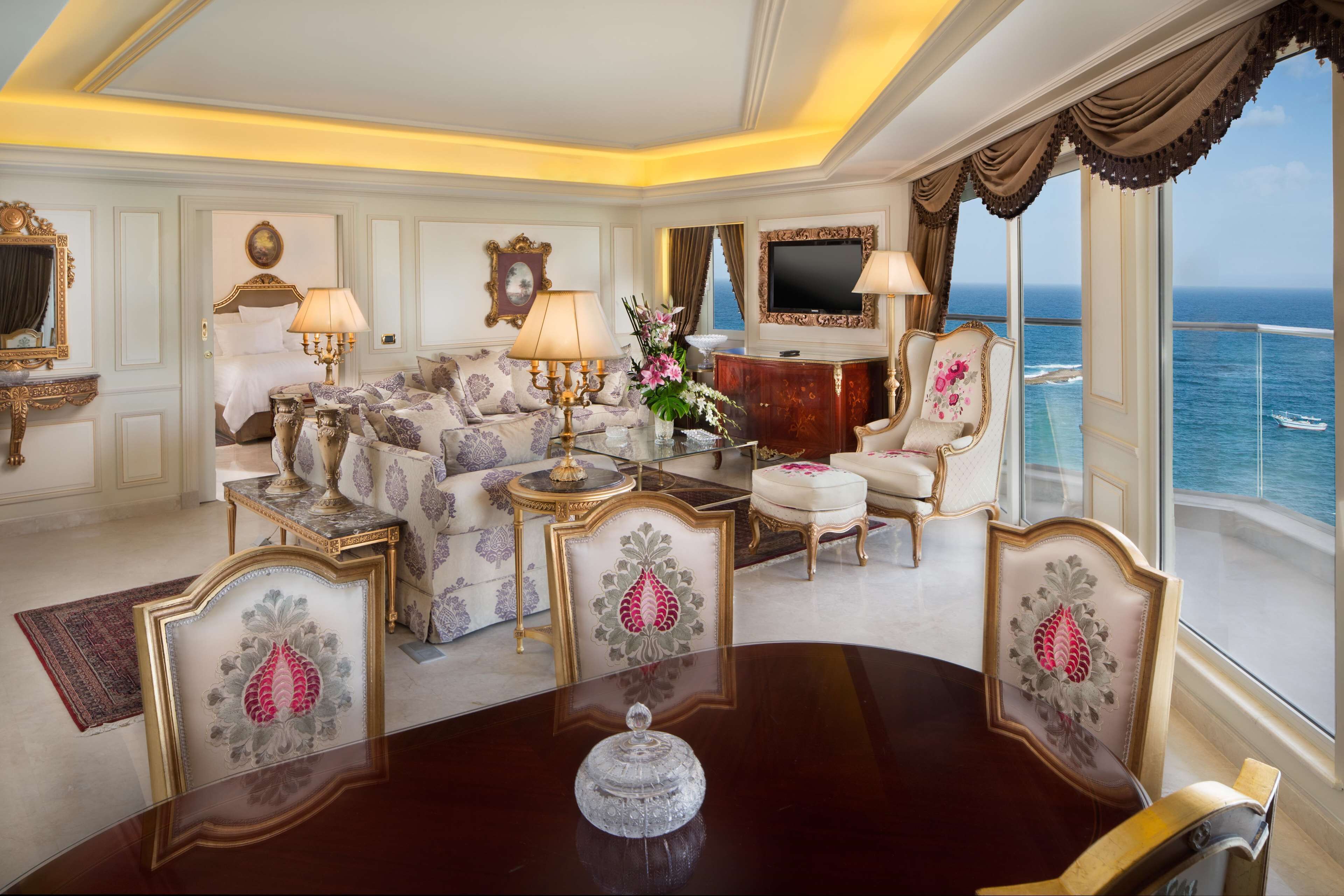 A luxurious room with a large window overlooking the ocean, a plush sofa set, a dining table with a centerpiece, and elegant decor.