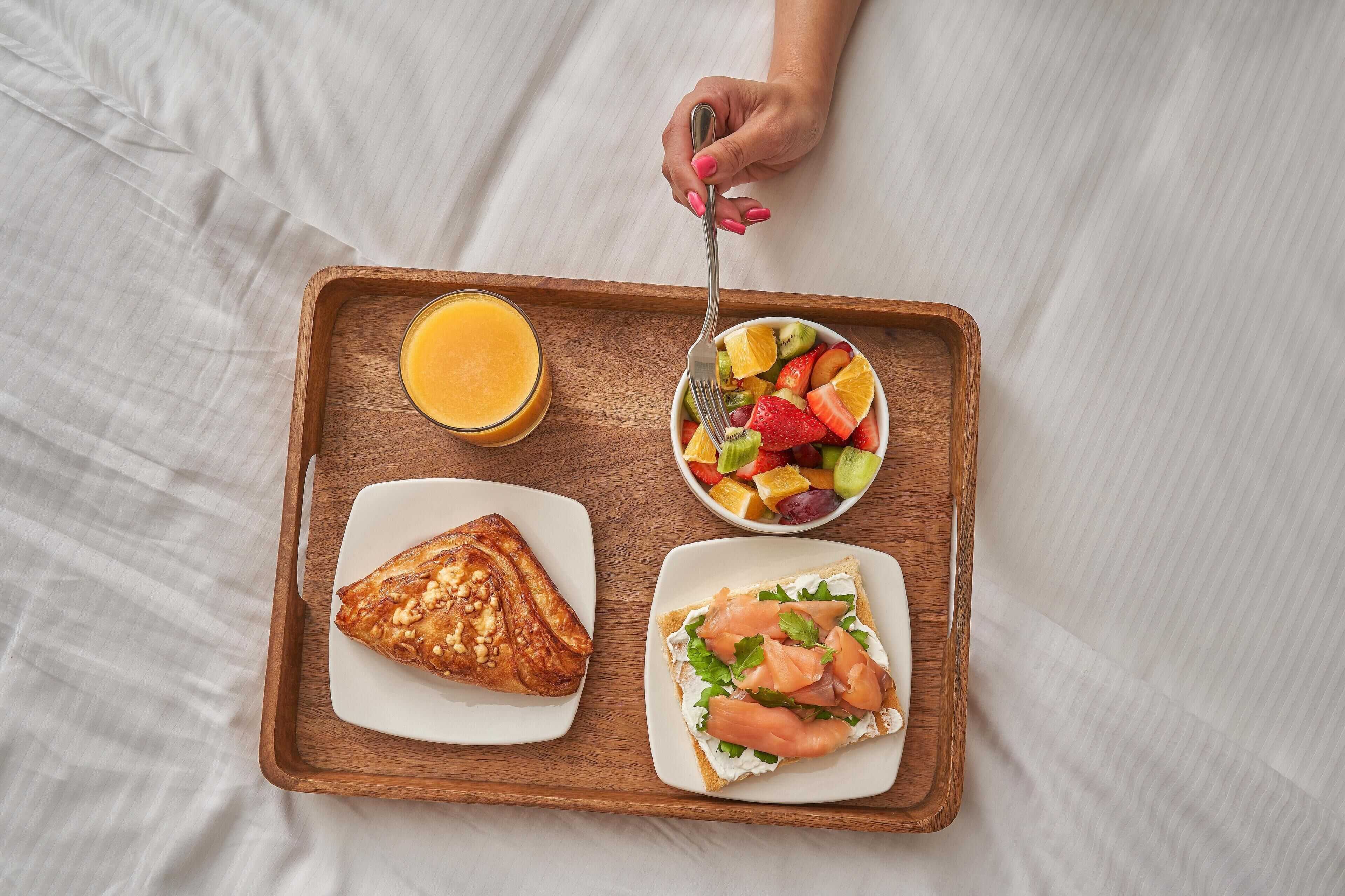 A breakfast tray with a fruit salad, a sandwich, a glass of orange juice, and a pastry on a wooden tray.