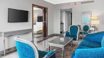 Habitación de hotel con una televisión de pantalla plana, una zona de comedor con mesa y sillas, un espejo y una cama al fondo.