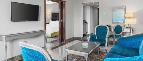 A hotel room with a flat-screen TV, a dining area with a table and chairs, a mirror, and a bed in the background.