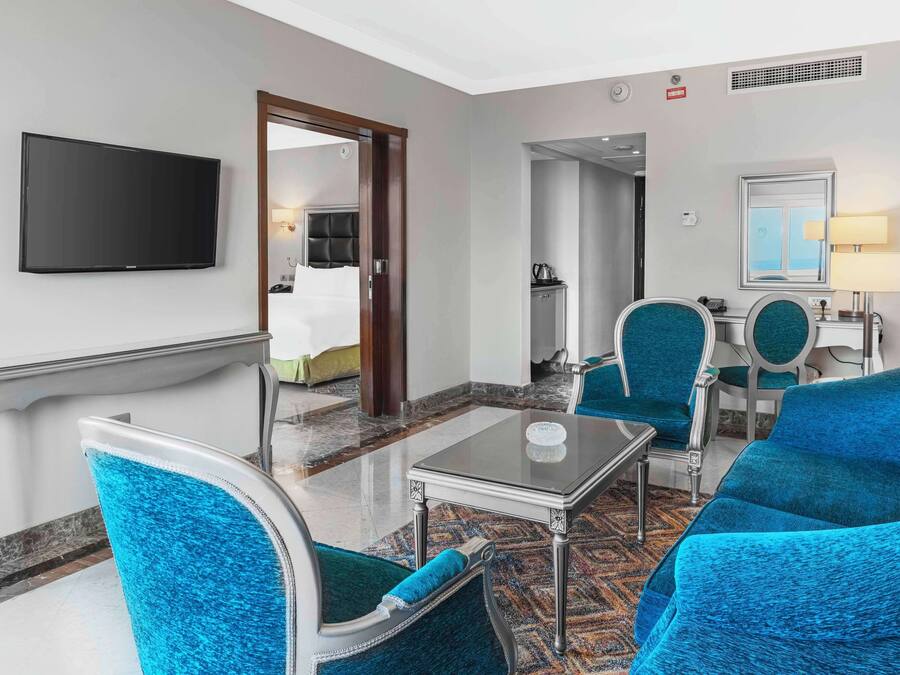 Habitación de hotel con una televisión de pantalla plana, una zona de comedor con mesa y sillas, un espejo y una cama al fondo.