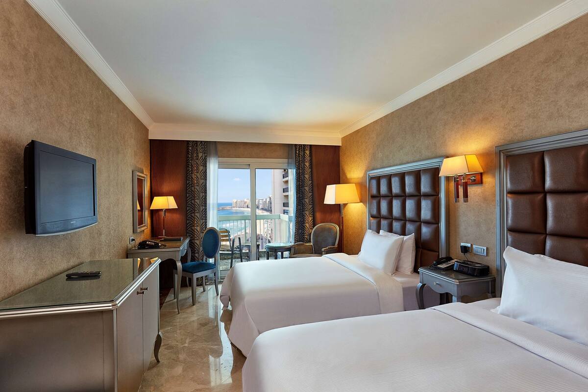 A hotel room with two beds, a desk, a chair, a TV, and a balcony with a view.