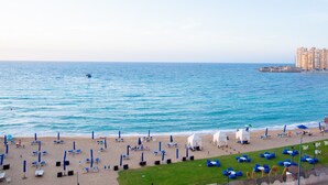 A beach with blue umbrellas and lounge chairs, a sandy shore, and a view of the ocean. - Hilton Alexandria Corniche (Alexandria)