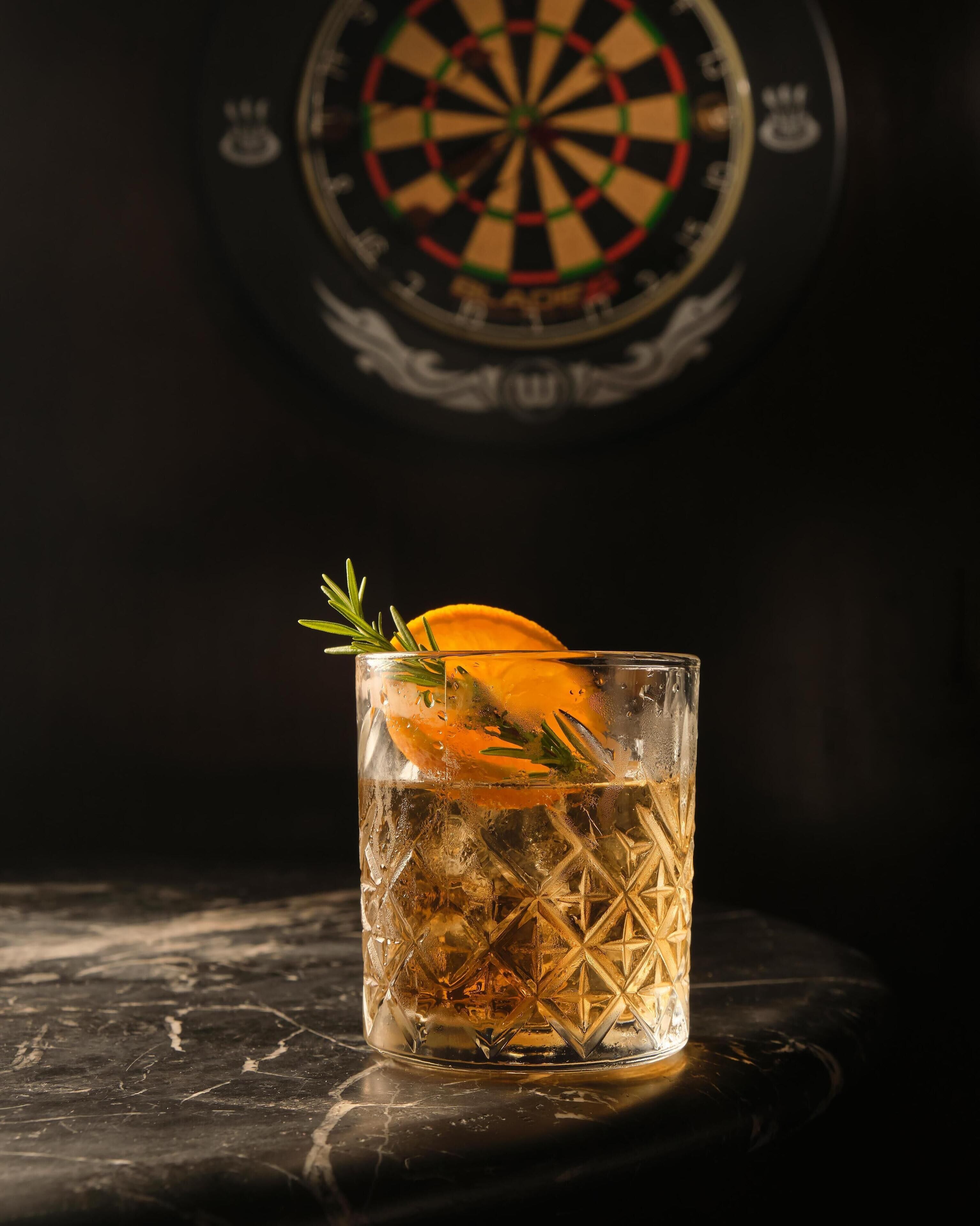 Un verre agrémenté d’une garniture, de glaçons, sur un fond sombre avec un jeu de fléchettes.