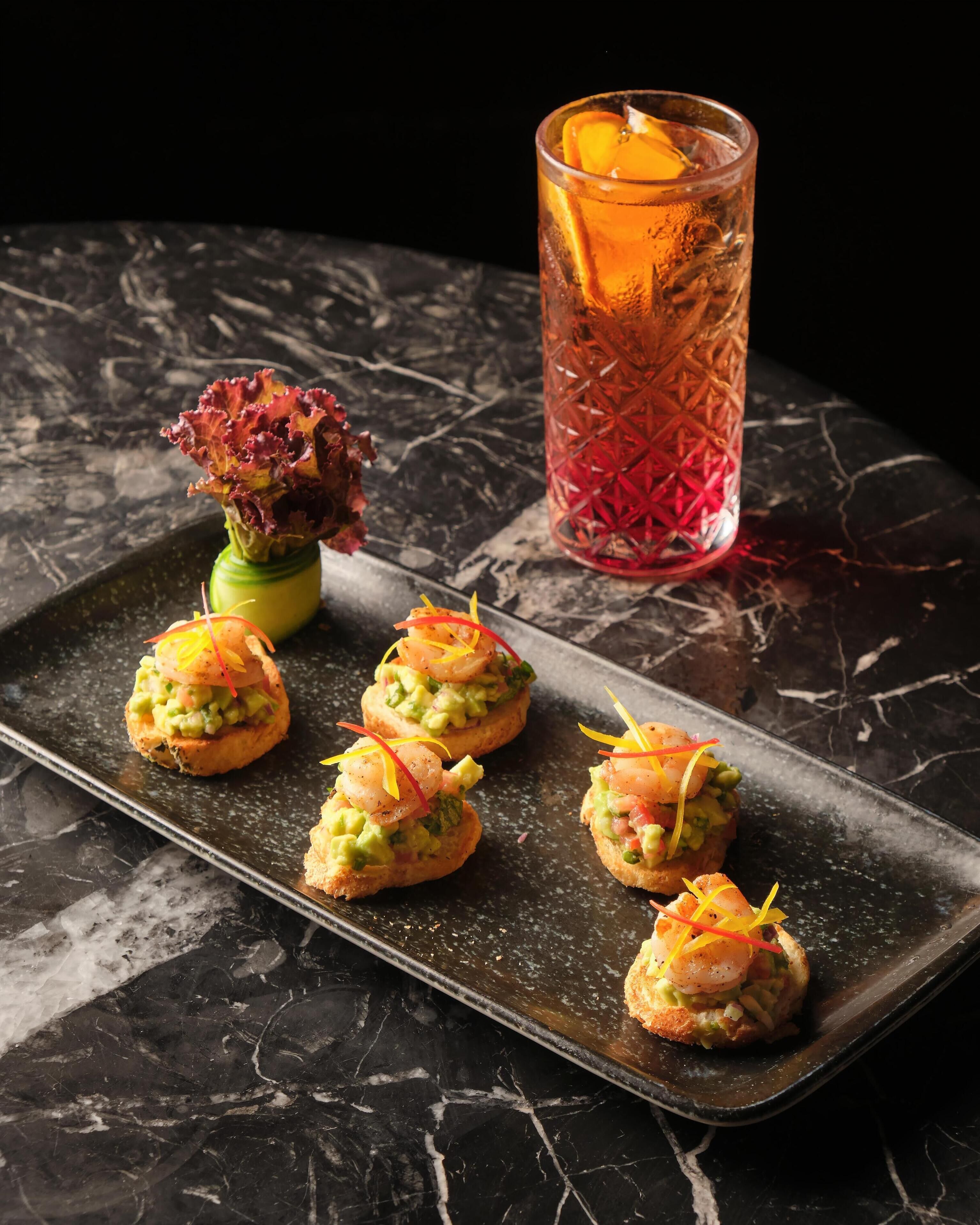 A tray of appetizers with shrimp and avocado on a dark marble table, a glass with a red pattern, and a decorative flower.