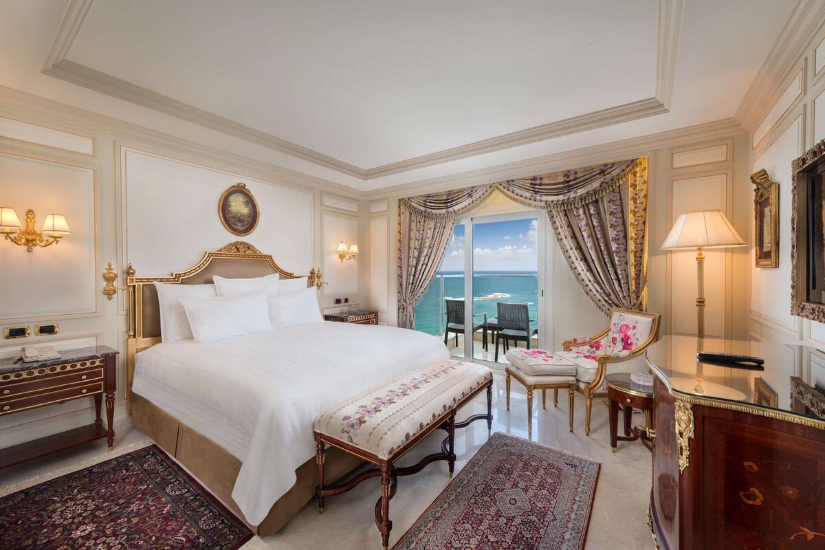 A luxurious bedroom with a large bed, a view of the sea, a sitting area with a chair and table, and elegant decor.