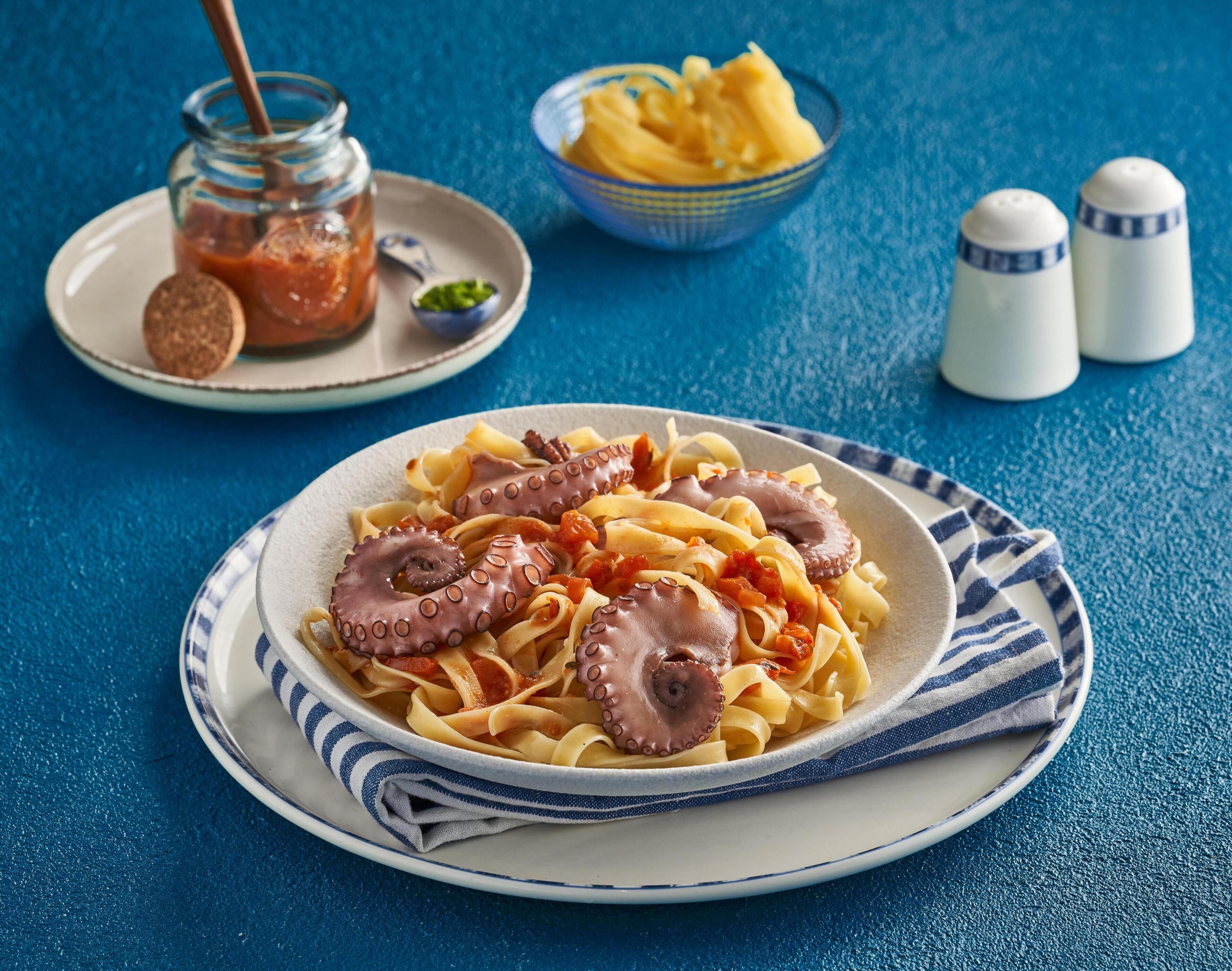 Une assiette de pâtes garnie de tentacules de poulpe, un bocal de sauce et un bol de fruits jaunes sur une table bleue.