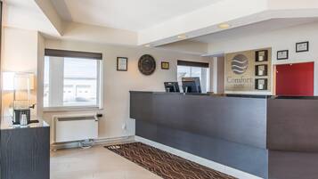 Zona de recepción de un hotel con mostrador, un reloj, diplomas enmarcados y una puerta roja.