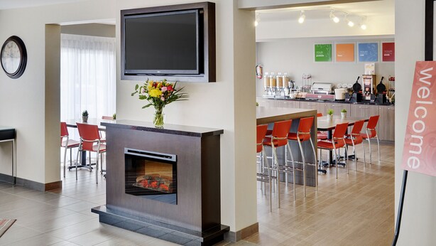 Un vestíbulo de hotel moderno con chimenea, sillas rojas, una zona de cocina y un cartel de bienvenida.