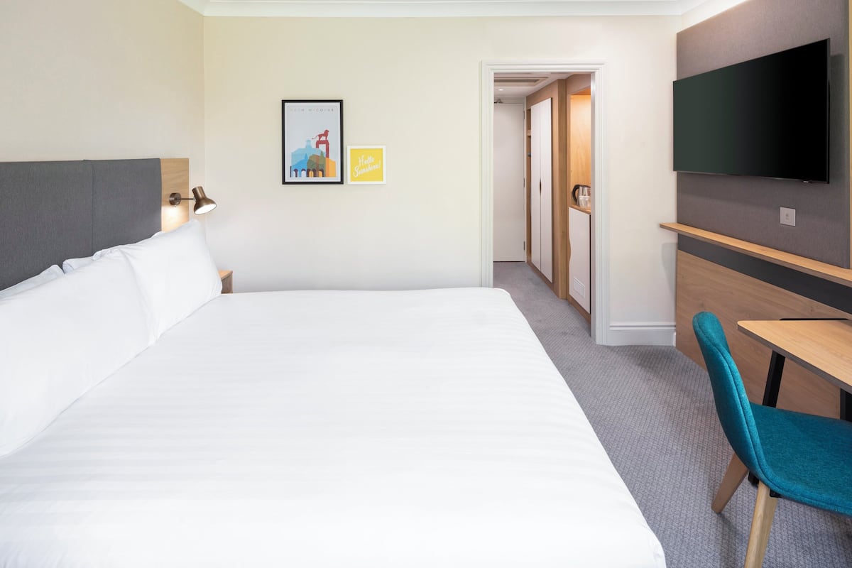 A hotel room with a large bed, a desk, a chair, a TV, and a door leading to another room.
