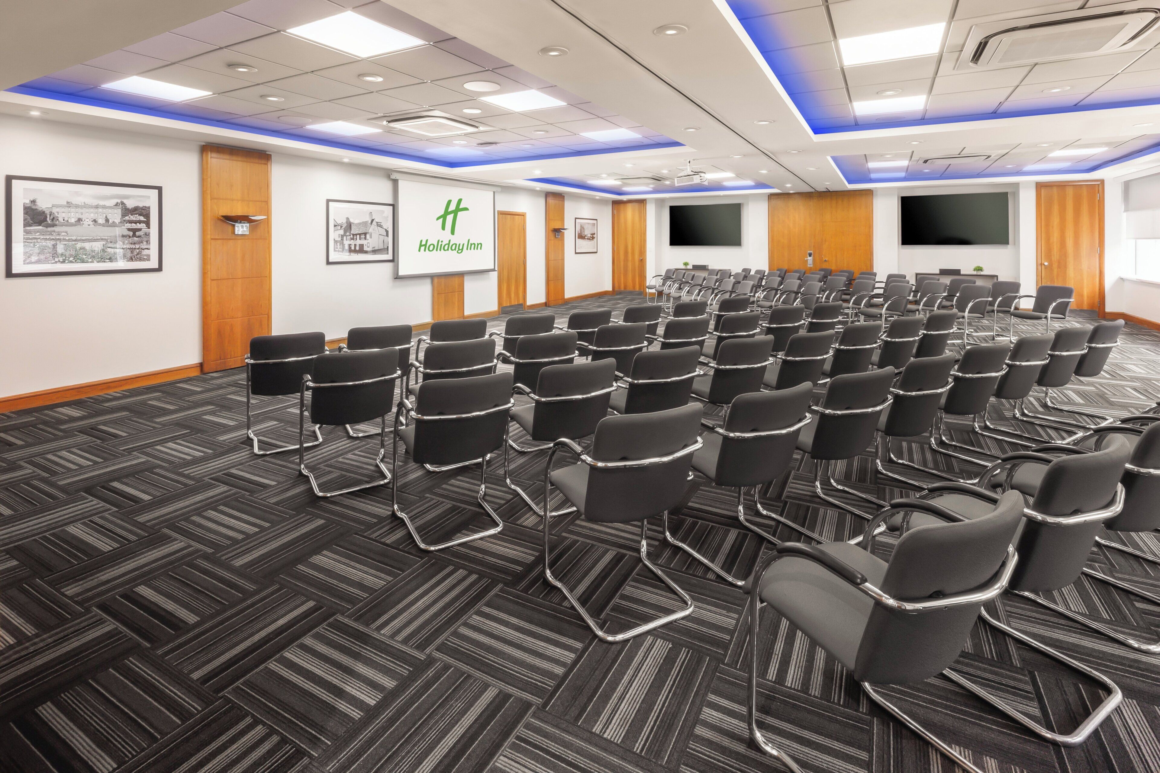 A conference room with rows of chairs, a large screen, and a Holiday Inn sign.