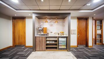 Een hotellobby met een receptiebalie, een koffieapparaat, een koelkast vol drankjes en een houten deur aan de rechterkant.