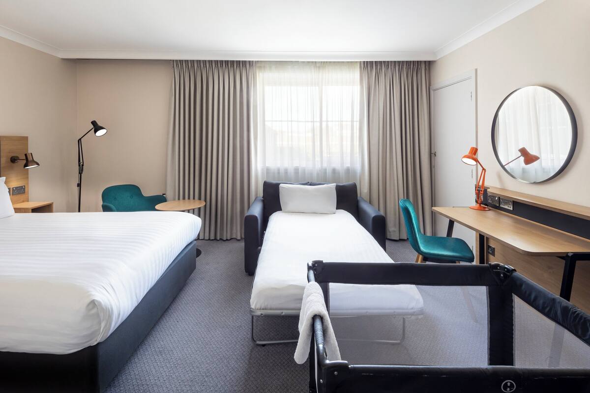 A hotel room with two beds, a desk, a chair, a small table, a mirror, and a window with curtains.