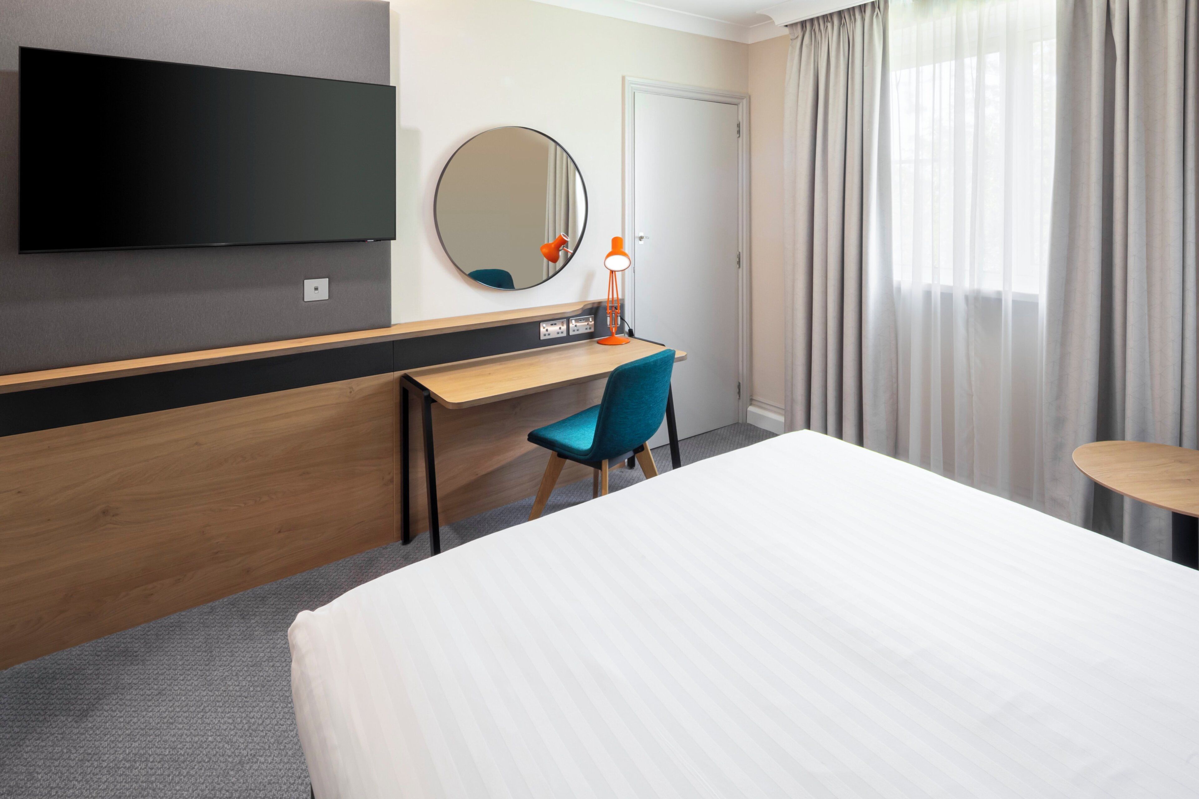 A hotel room with a bed, a desk, a chair, a TV mounted on the wall, a round mirror, and a window with curtains.