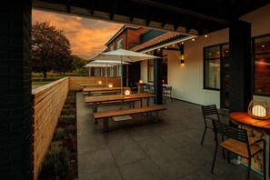 Eine Terrasse im Freien mit Holzbänken und -tischen, einer Ziegelwand und Blick auf Baum und Himmel.